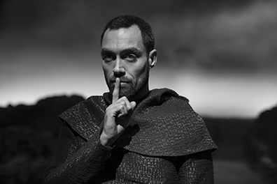 Alex Hassell as the Thane of Ross, in chainmail shirt and cloaked shoulder with his finger up to his lips in a "silence" gesture (black and white photograph).