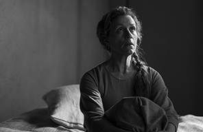 Francis McDormand as Lady Macbeth sitting on a bed with with her knees bent up to her torso, wearing night dress, pillow in the background (black and white photograpm).