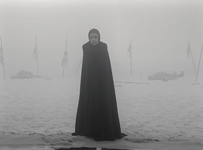 Kathryn Hunter as a witch in black hood and ground-lengh robe on a foggy beach with in the backgroun bodies lying on the sand and six flags stuck into the ground (black and white photograph).